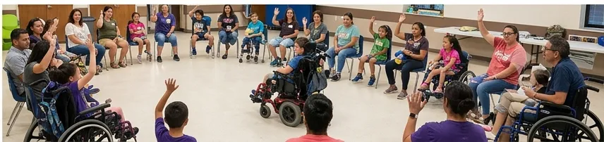 A large circle of children and volunteers seated together in a community hall, many with hands raised